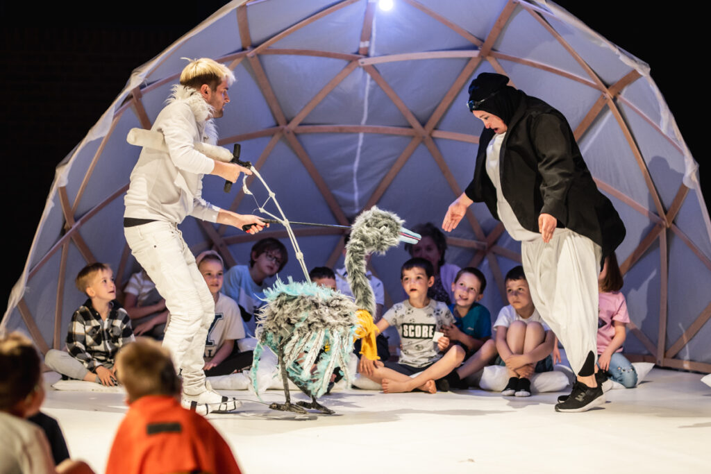 Na środku sceny aktor prowadzi kukiełkę ptaka. Przed nim aktorka ubrana w strój pingwina. Za nimi w połówce wielkiego igloo siedzą dzieci.