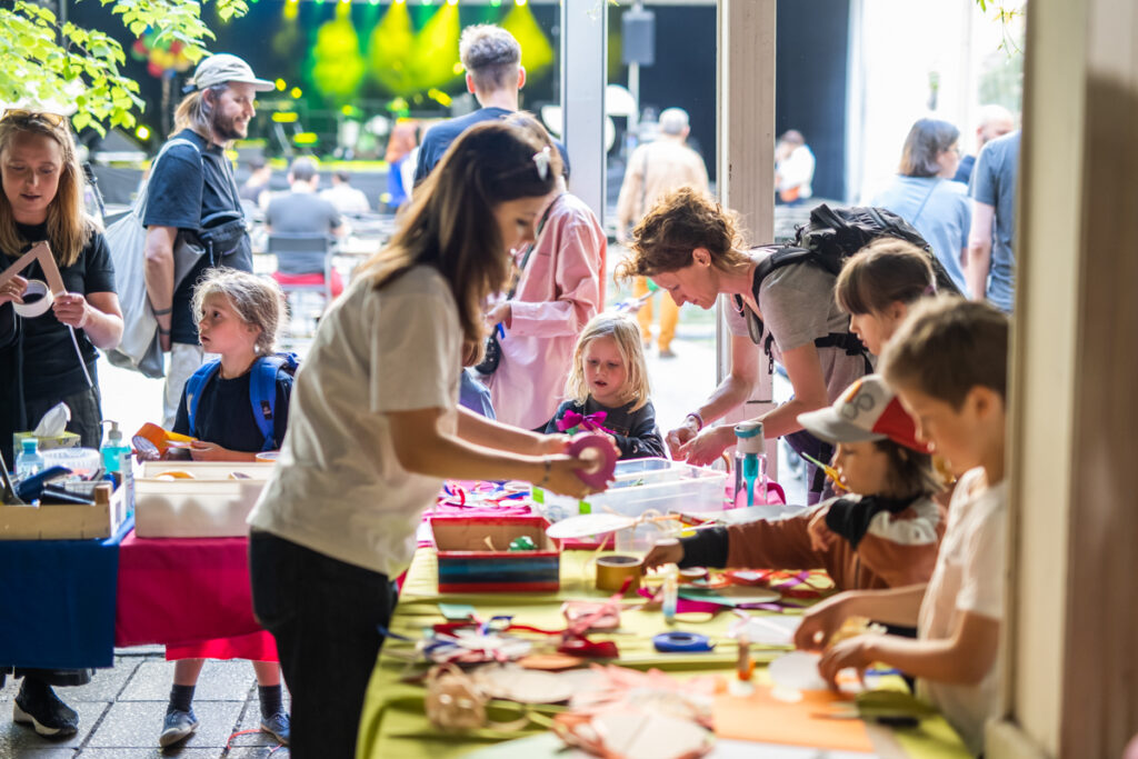 Grupa dzieci i dorosłych przy stołach zastawionych materiałami i przyborami plastycznymi.