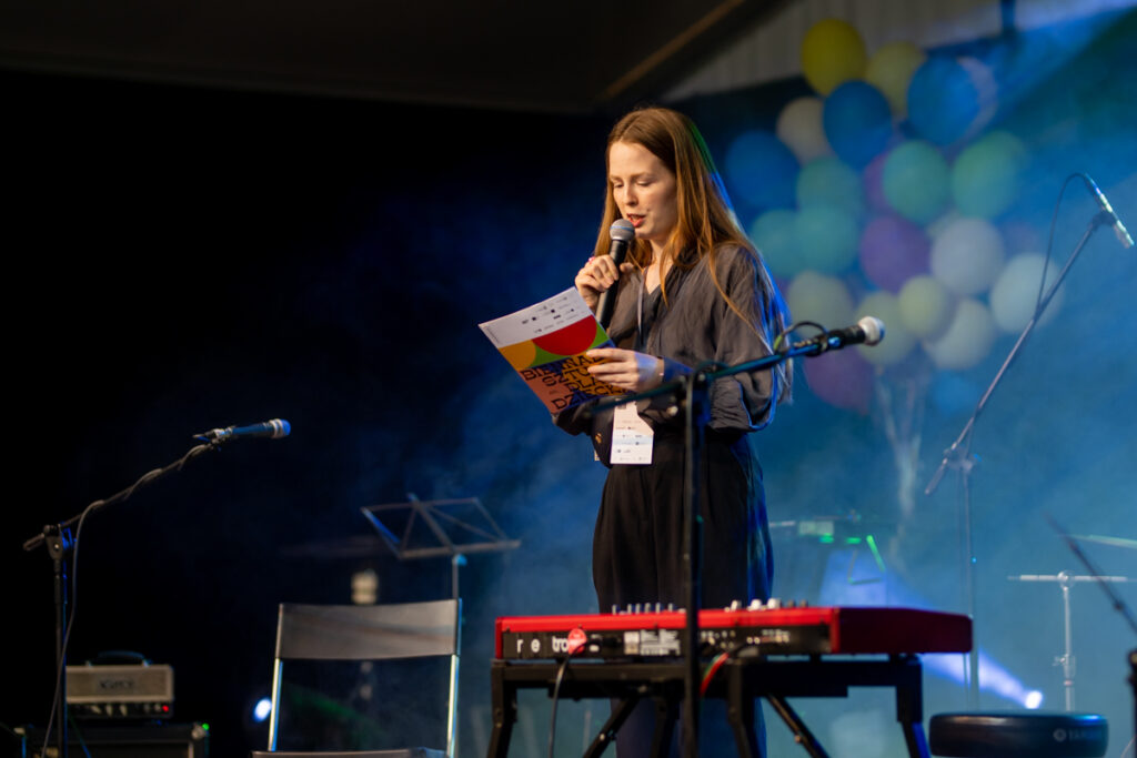 Kobieta stoi na scenie pośród instrumentów muzycznych. Czyta coś do mikrofonu.