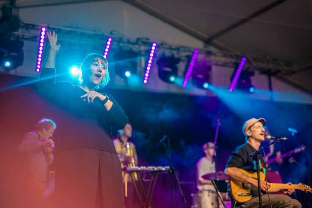 Na scenie grupa muzyków. Po lewej kobieta używająca języka migowego.