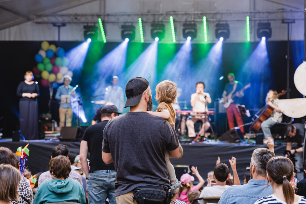 W tle grupa muzyków na scenie. Z przodu widownia. Mężczyzna trzyma dziecko na rękach.