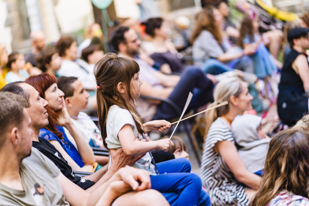 Na widowni grupa dzieci i dorosłych. Na środku dziewczynka siedzi na kolanach mężczyzny. Ma w rękach drewniane pałeczki.