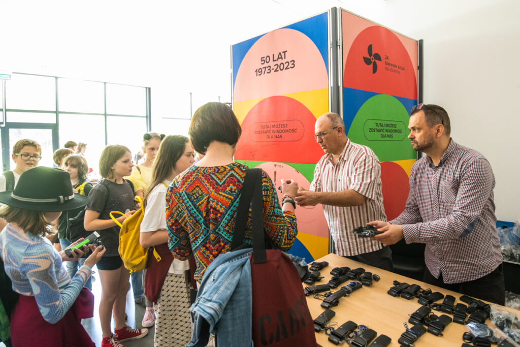 Po prawej dwóch mężczyzn rozdaje aparaty do tłumaczenia. Obok nich grupa dzieci i dorosłych.