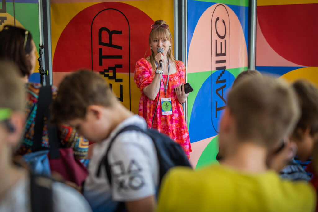 Kobieta przemawia do grupy dzieci i dorosłych ustawionych tyłem.