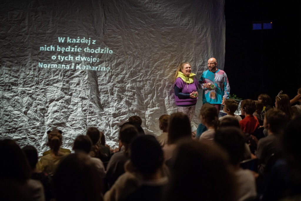 Na scenie dwójka aktorów. Za nimi płachta, na której wyświetla się napis: w każdej z nich będzie chodziło o tych dwoje: Nornama i Katherine.