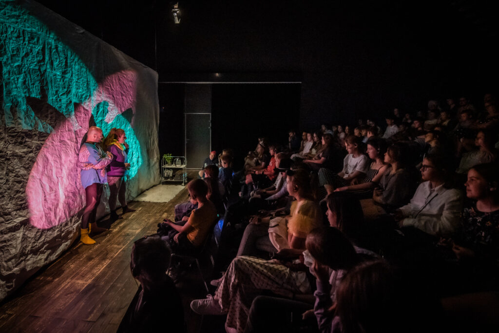 Po prawej widownia w półmroku. Po lewej scena z dójką aktorów. Za nimi unosi się płachta, na którą pada zielne i różowe światło.