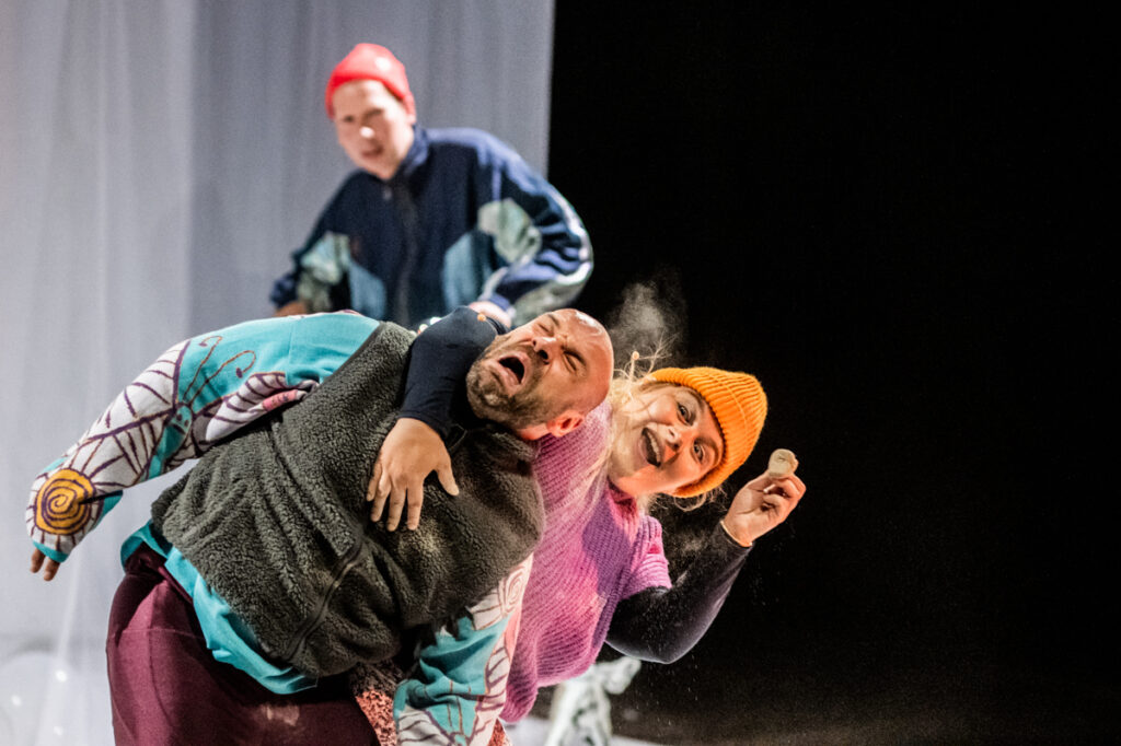 Na scenie trójka aktorów. Aktorka trzyma wygięta na bok aktora, który ma wykrzywioną twarz. W tle spogląda na nic inny aktor.