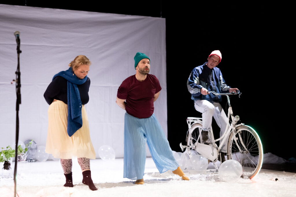 Trójka aktorów na scenie. Jeden z nich siedzi na rowerze. Obok niego stoją aktor i aktorka, którzy grzebią stopą w rozrzuconym po scenie styropianie.