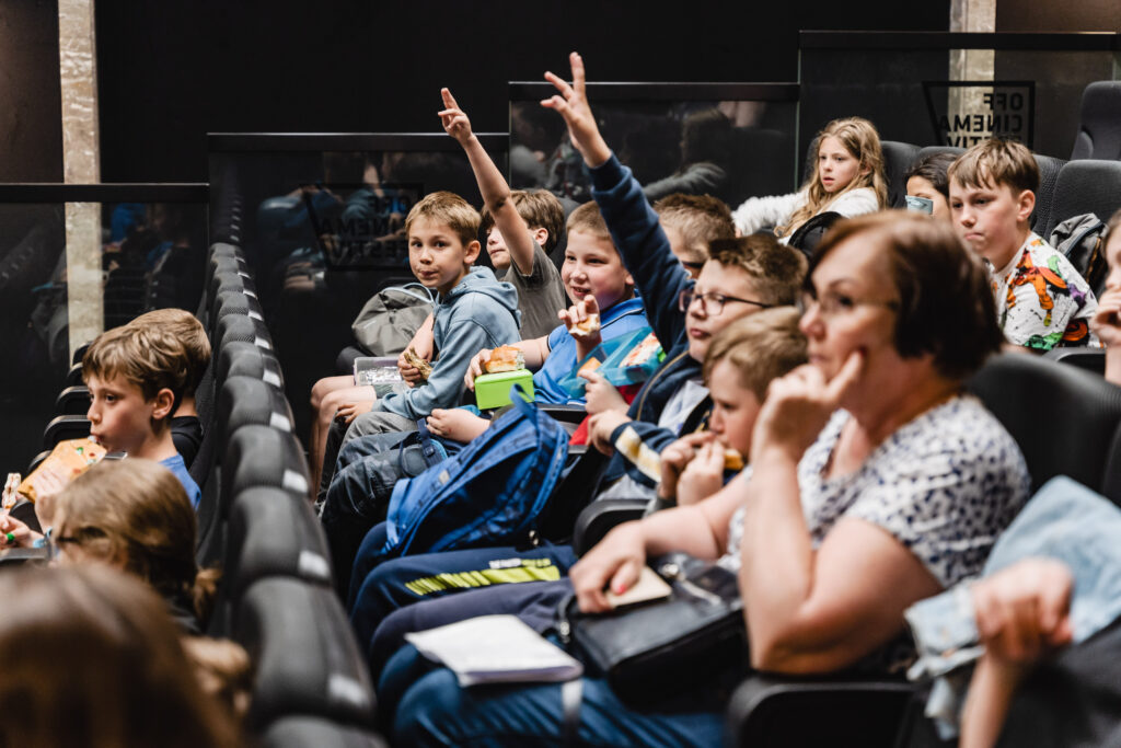 Na widowni grupa dzieci i dorosłych. Kilkoro dzieci unosi ręce do góry.
