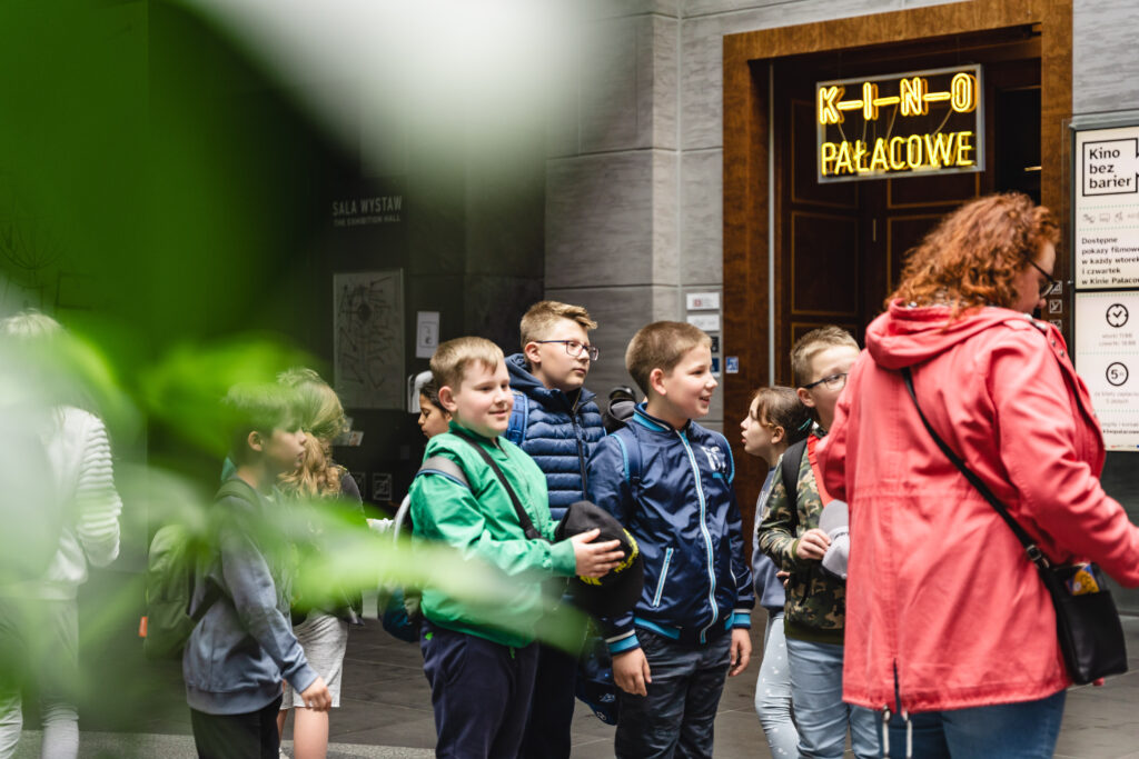 Grupa dzieci i dorosłych stoi w holu poznańskiego Zamku. W tle widać napis: Kino Pałacowe.