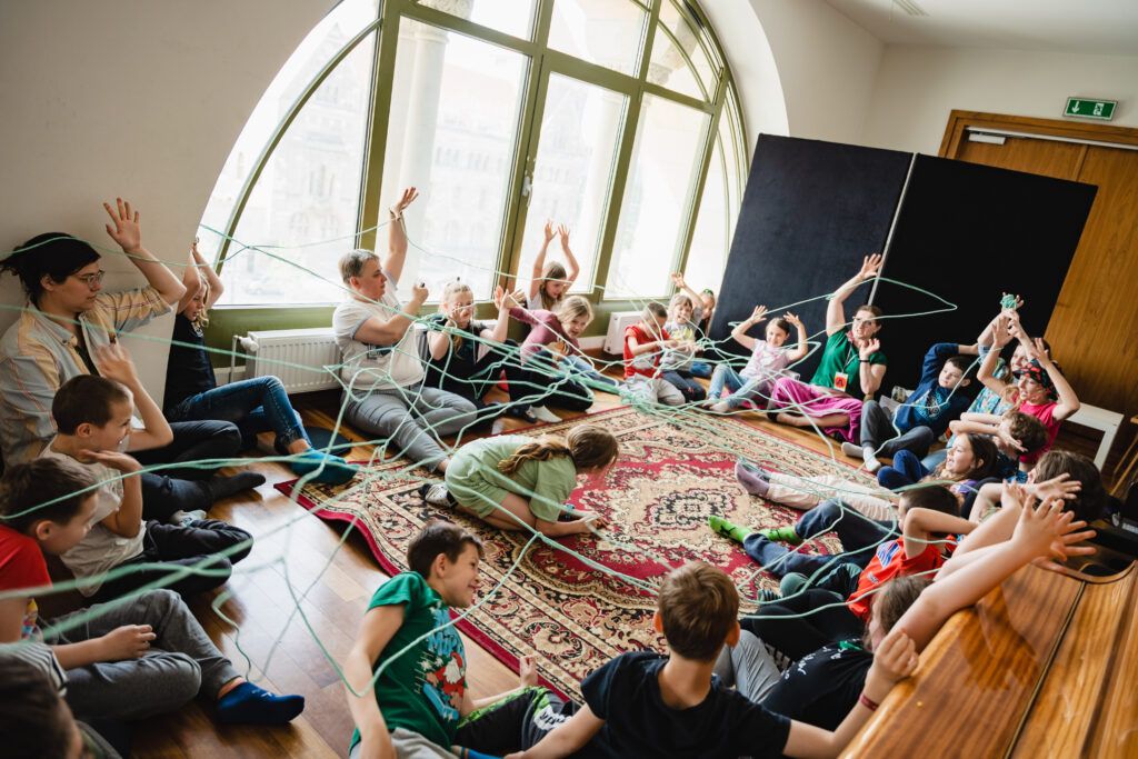 W sali grupa dzieci i dorosłych siedzi na około. Nad nimi unosi się pajęczyna zrobiona z nitek.