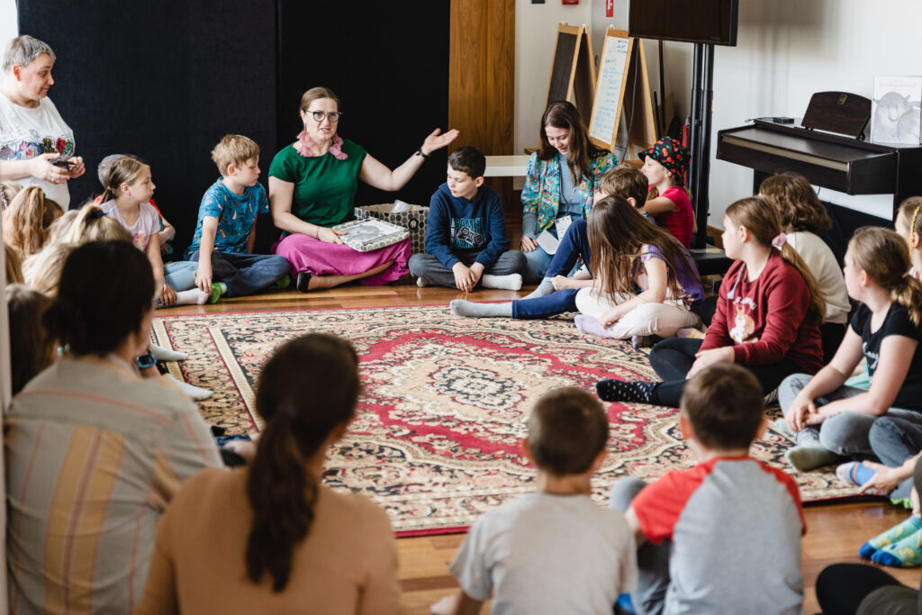 W sali w kręgu siedzi grupa dzieci i dorosłych. Na środku kobieta w okularach opowiada.