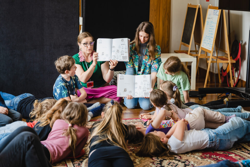 Na podłodze leży grupa dzieci. Przed nimi dwie kobiety prezentują dwie książki.