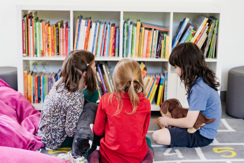 Grupa dzieci siedzi. Obok nich regał z książkami.
