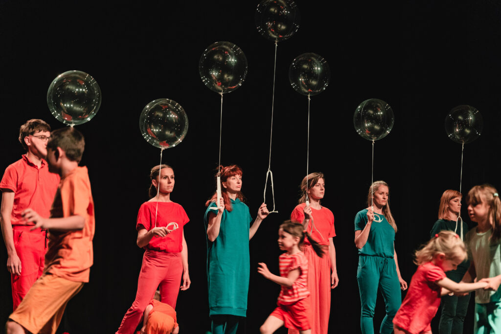 Na scenie grupa osób ustawione w rzędzie. Trzymają w rękach przeźroczyste balony. Przed nimi biegają dzieci.