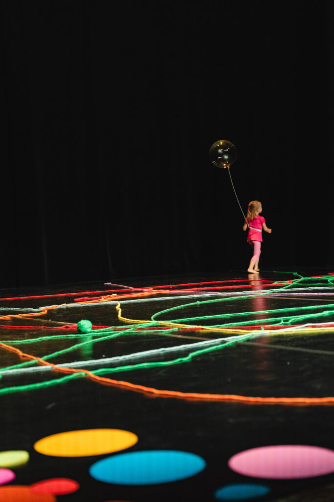 Na scenie samotna dziewczynka z przeźroczystym balonem. Na podłodze rozłożone są kolorowe nitki.