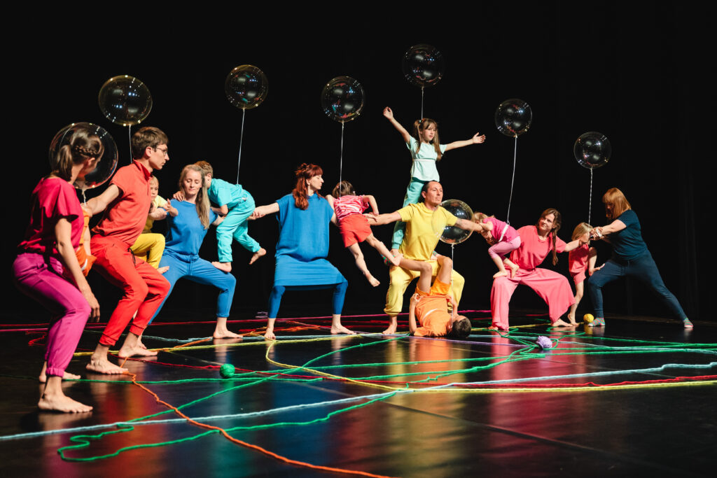 Na scenie grupa dzieci i dorosłych. Dorośli stoją w rozkroku trzymając się za ręce. Dzieci wspinają się po nich lub wiszą na ich ramionach.