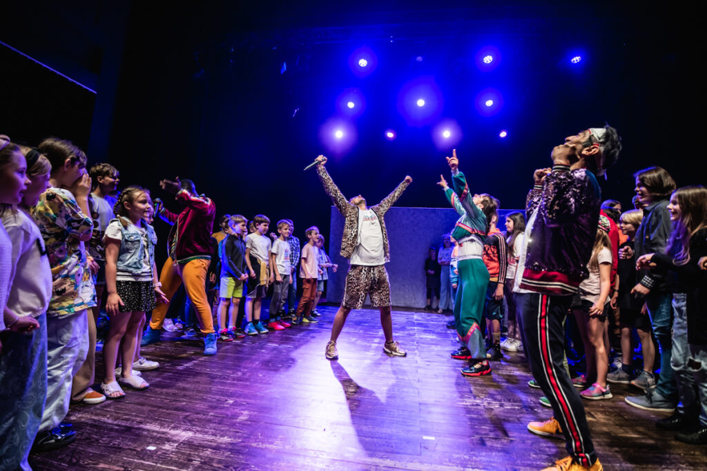 Dwójka aktorów na środku sceny z uniesionymi rękoma. Pośród tłumu młodzieży.