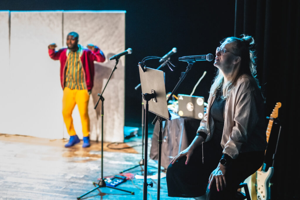 Po prawej piosenkarka śpiewa. W tle po lewej aktor tańczy na scenie.