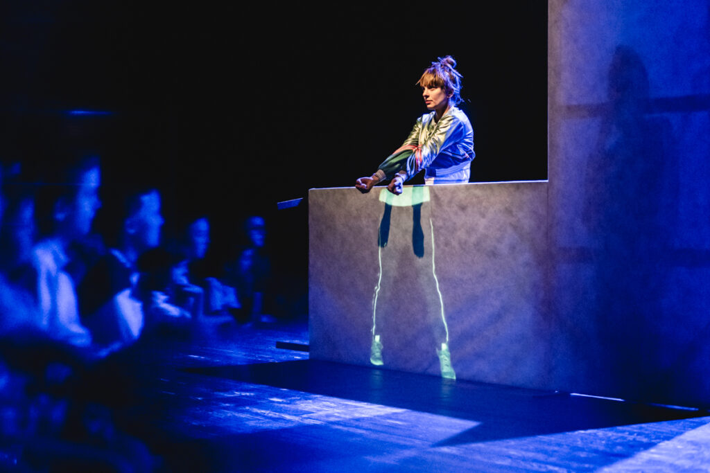 Aktorka stoi za ekranem, zasłaniającą ją do połowy i tańczy.