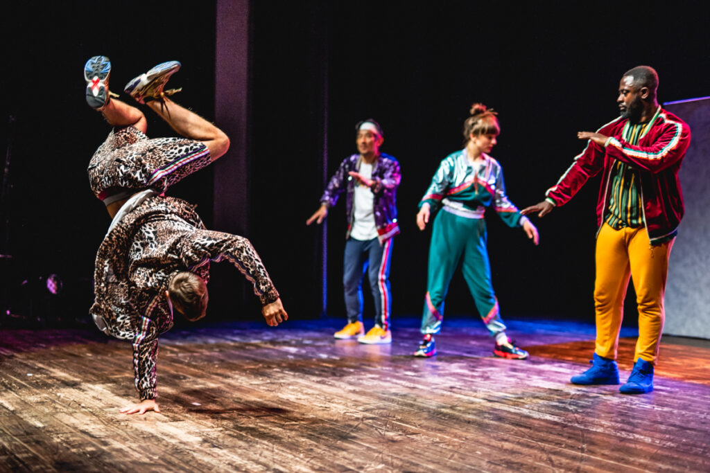 Grupa aktorów tańczy na scenie. Aktor po lewej stornie stojąc na jednej ręce wykonuje figurę taneczną.