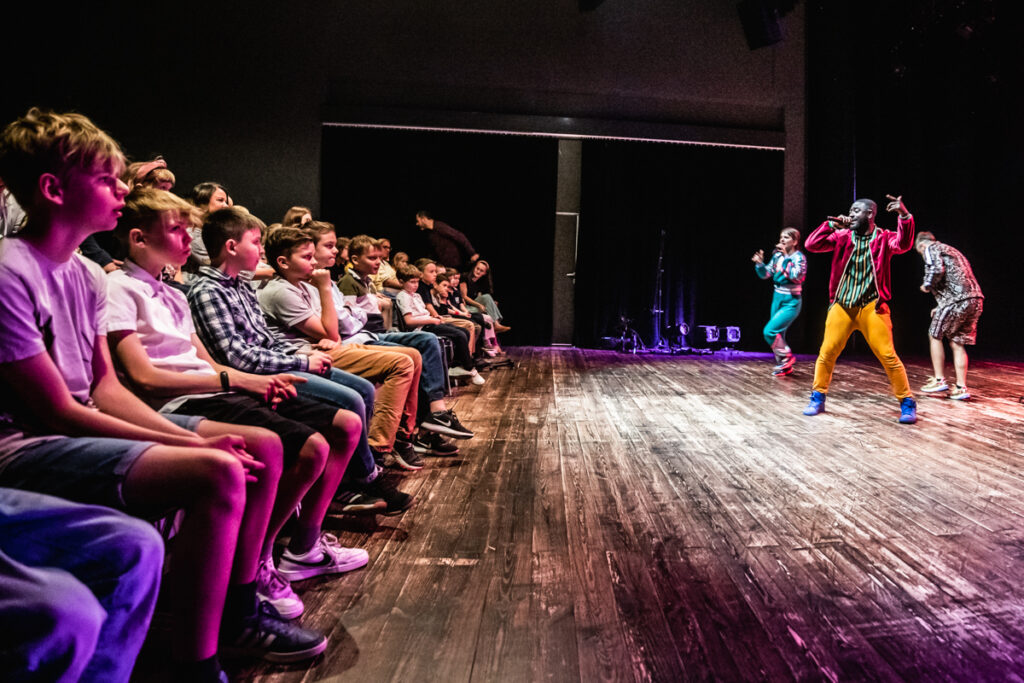 Po lewej stronie dzieci zgromadzone na widowni. Po prawej aktorzy tańczą na scenie.