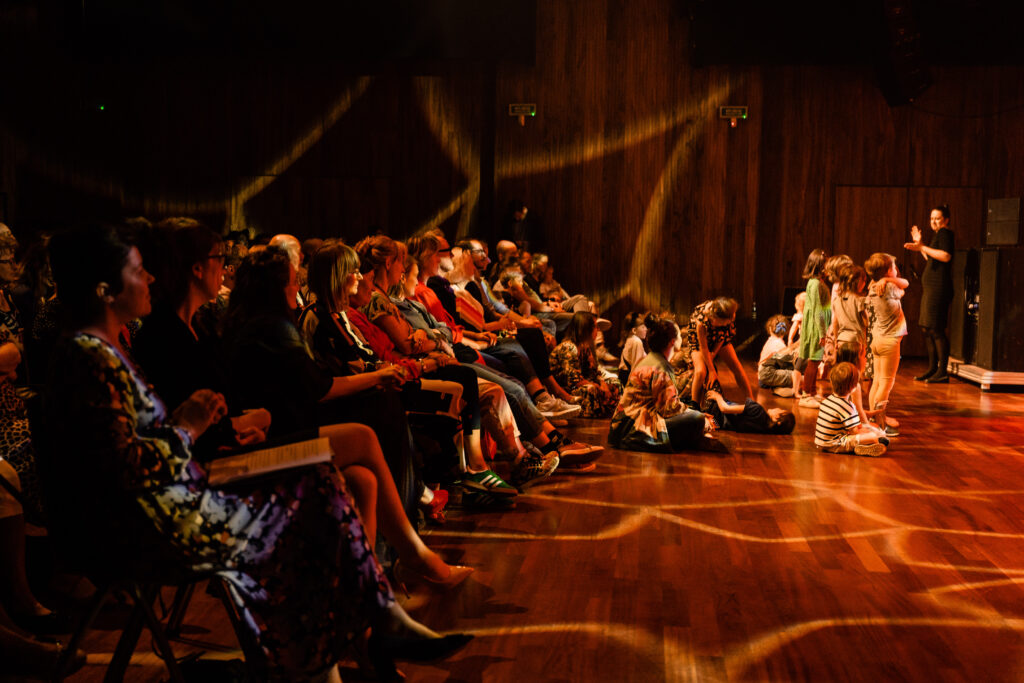 Osoby dziesiące na widowni. Grupka dzieci stoi z przodu. W tle kobieta używająca języka migowego.