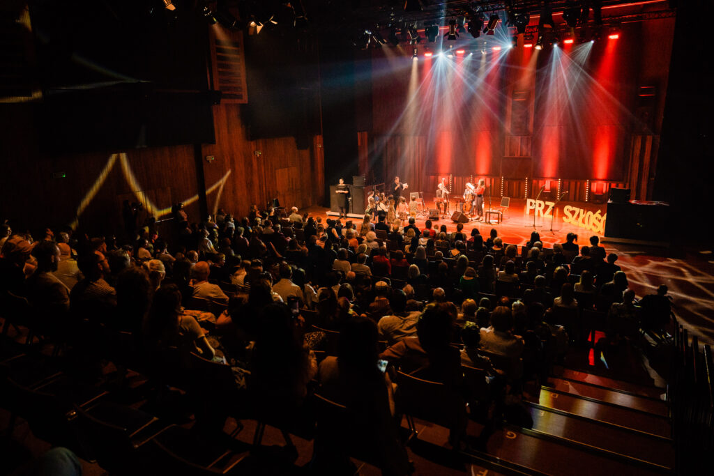 Na przedzie osoby siedziące na widowni. W tle grupa muzyków gra na scenie.