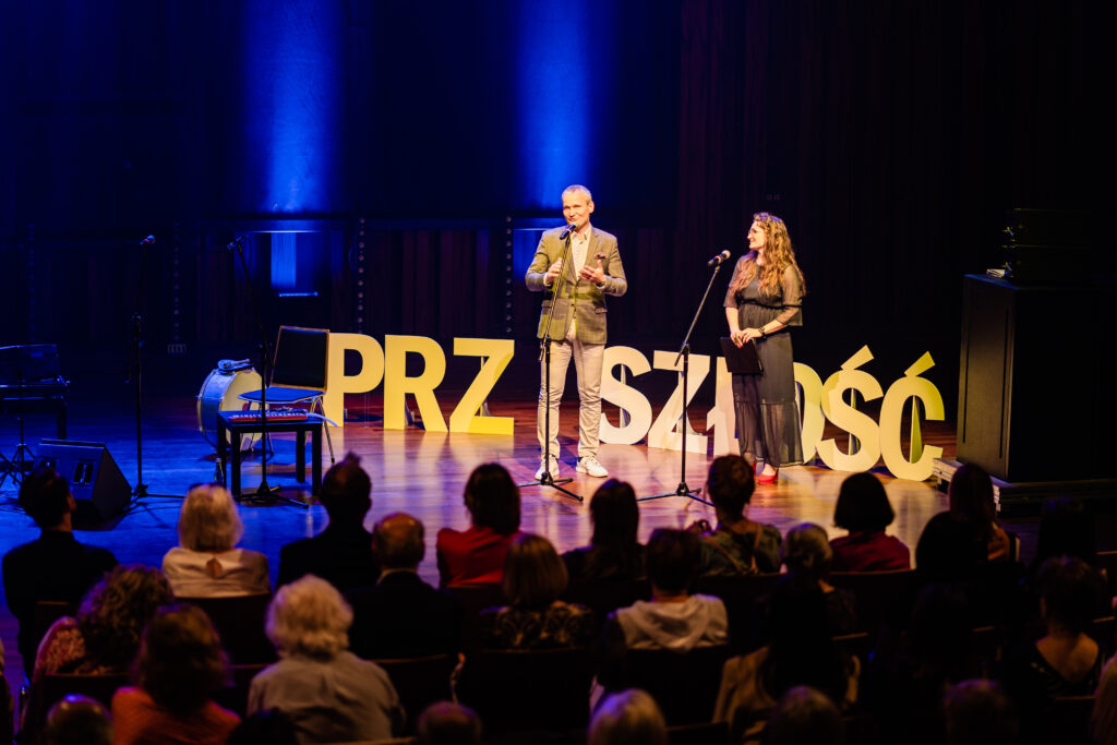 Dwoje prowadzących stoją przed osobami na widowni. Za nimi wielki napis: Przyszłość.