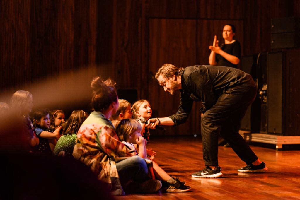 Mężczyzna podaje mikrofon dzieciom siędzącym na widowni. W tle kobieta używa języka migowego.