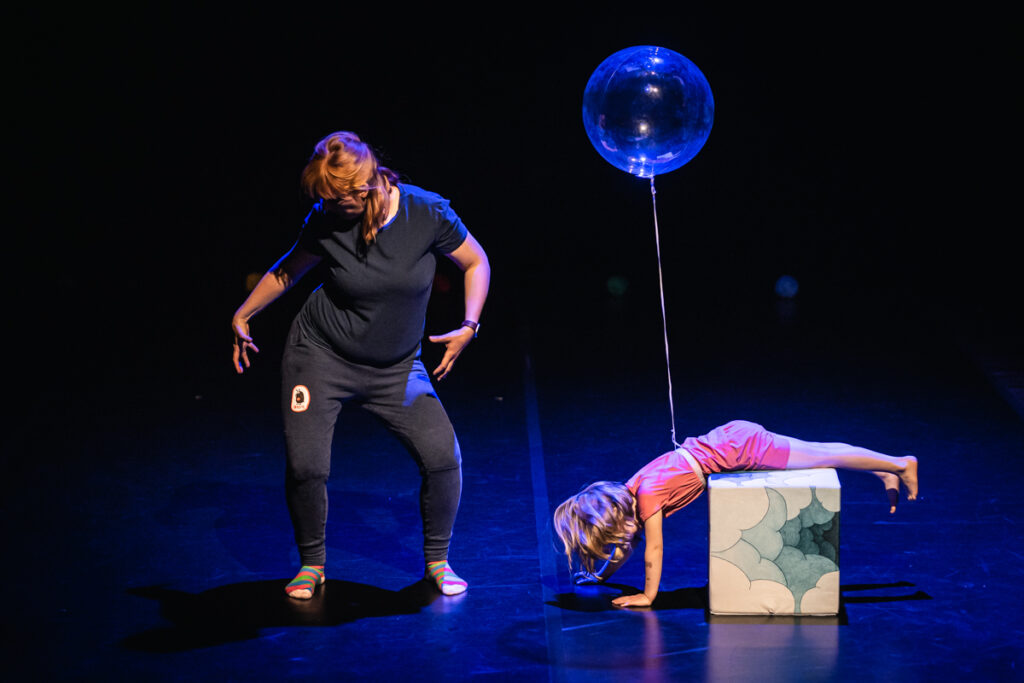 Na scenie kobieta tańczy. Obok niej dziewczynka nogami oparta o kolorowy sześcian.