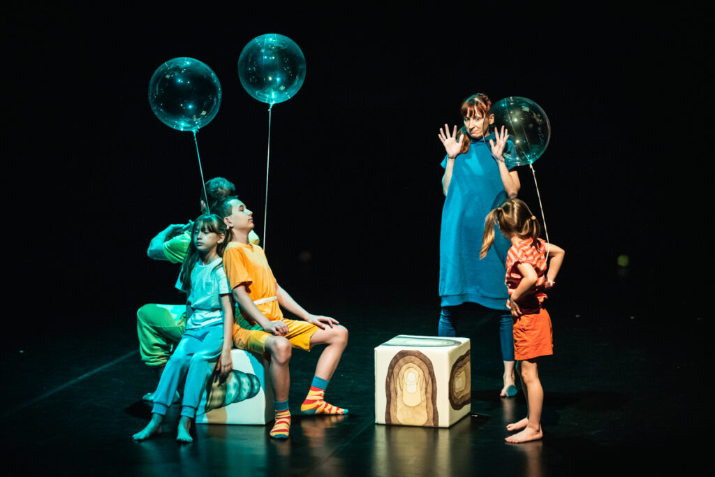 Na scenie grupa dzieci i dorosłych. Do dzieci przywiązane są przeźroczyste balony. Trójka osób siedzi na kolorowym sześcianie. Po prawej leży drugi taki sześcian. Za nim stoi kobieta z uniesionymi rękoma.
