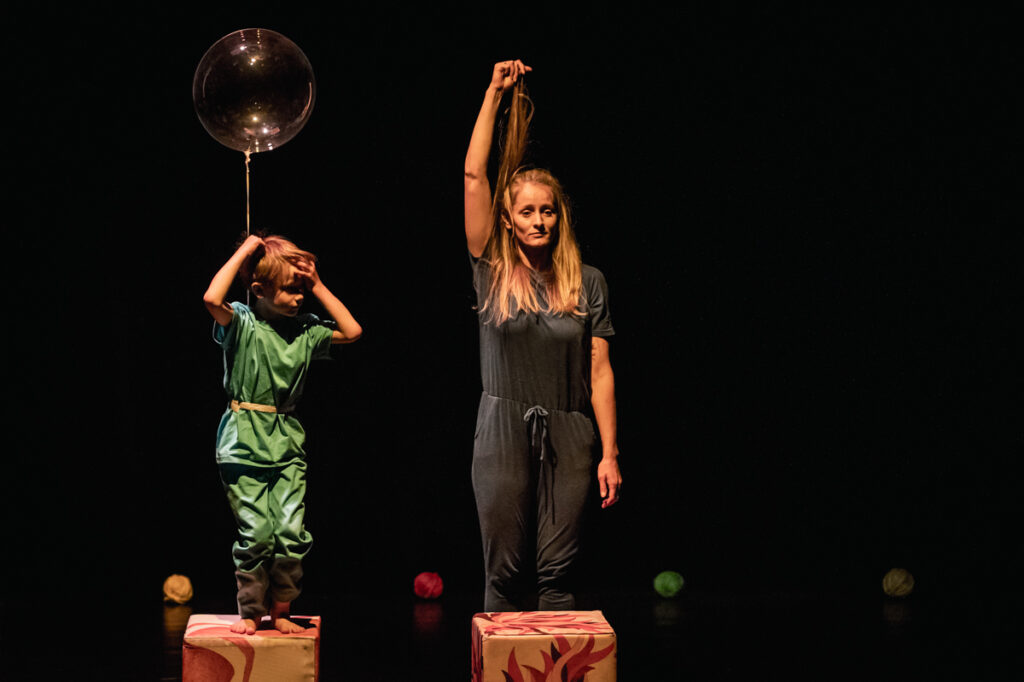 Na scenie kobiet wysoko uniesioną ręką, w której trzyma swoje włosy. Przed nią leży kolorowy sześcian. Na drugim takim sześcianie stoi dziecko, do którego przywiązany jest kolorowy balon.