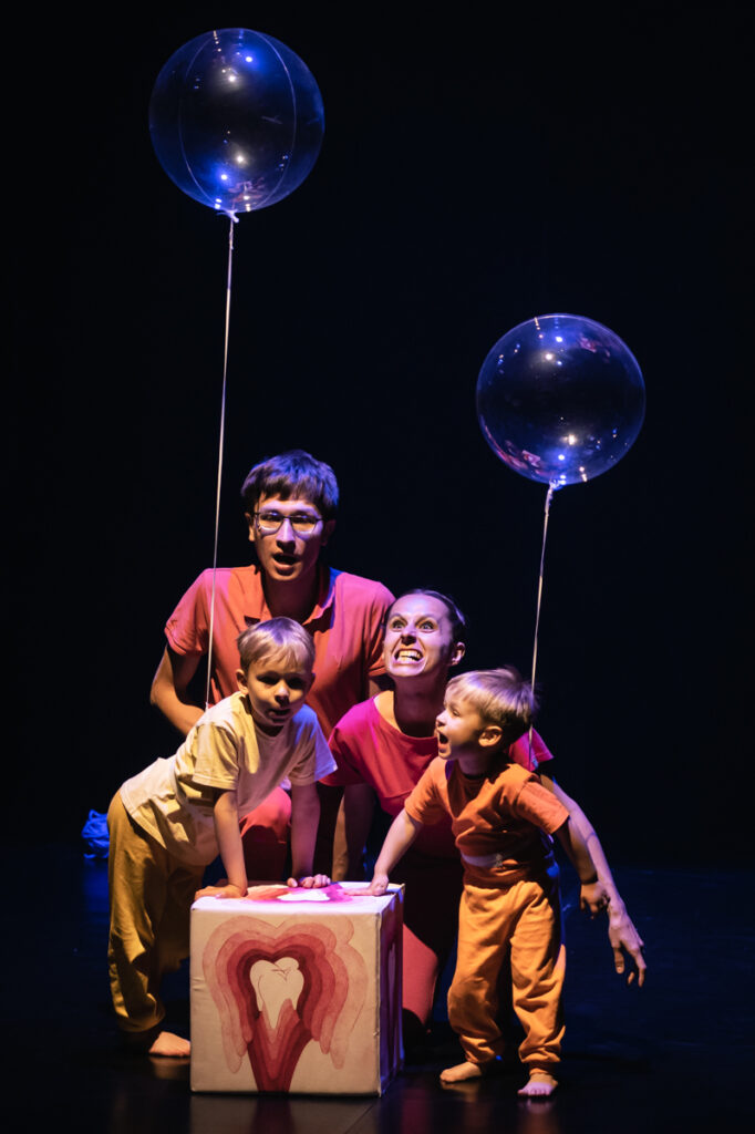 Na scenie grupa osób. Mężczyzna i kobieta siedzą z tylu. Kobieta ma wyszczerzone zęby. Na przedzie stoi dwóch chłopców opierających się o dużą stojącą kostkę. Do obu przywiązane są przeźroczyste balony.