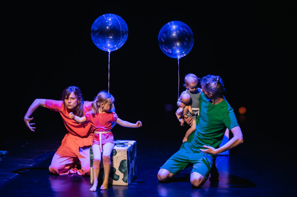 Po lewej kobieta z szeroko rozstawionymi rękoma. Obok niej na dużej kostce siedzi dziewczynka. Po prawej klęczący mężczyzna z dzieckiem na rękach. Nad nimi unoszą się dwa przeźroczyste balony.