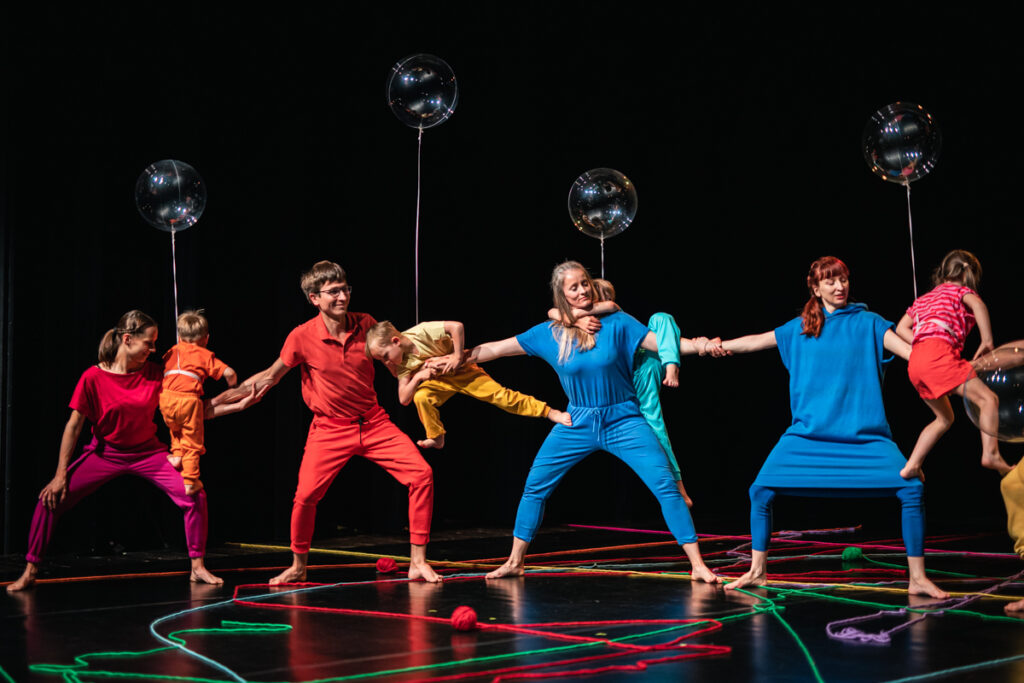 Grupa dorosłych stoi w rozkroku na scenie. Trzyma się za ręce. Na rękach widzą dzieci, do których przypięte są przeźroczyste balony.