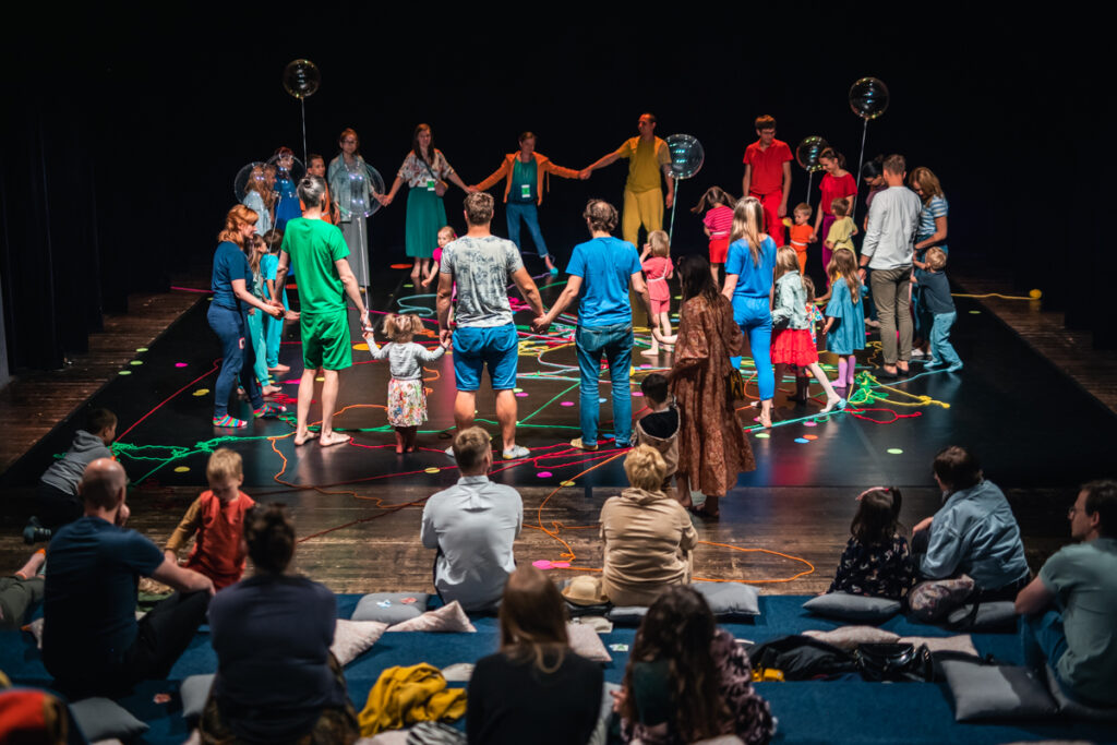 Z przodu widownia. Osoby siedzą na poduszkach. W tle scena, na które grupa dzieci i dorosłych ustawiona jest w kręgu . Na podłodze leżą kolorowe sznury.