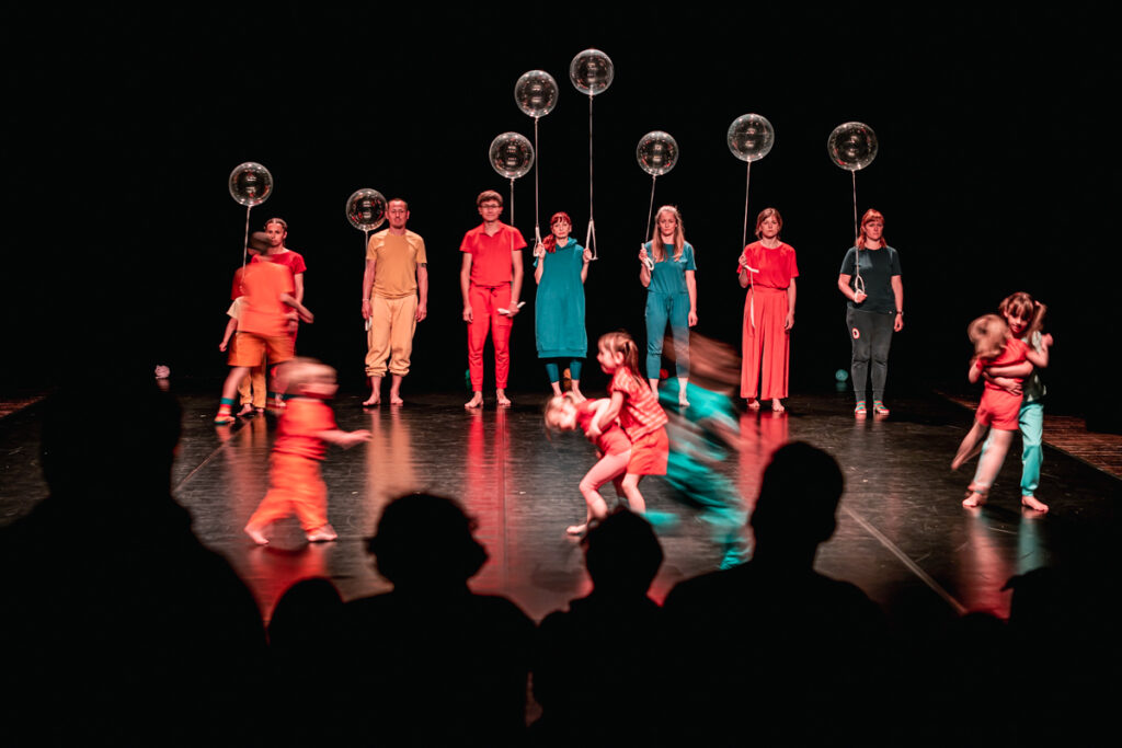Na scenie stoi grupa aktorów. Trzymają w rękach przeźroczyste balony. Po scenie biegają dzieci.