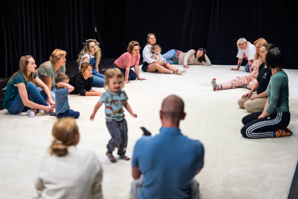 Na scenie grupa dzieci i dorosłych siedzi lub leży w kręgu. Pośrodku mała dziewczynka idzie w stronę siedzącego z przodu mężczyzny.