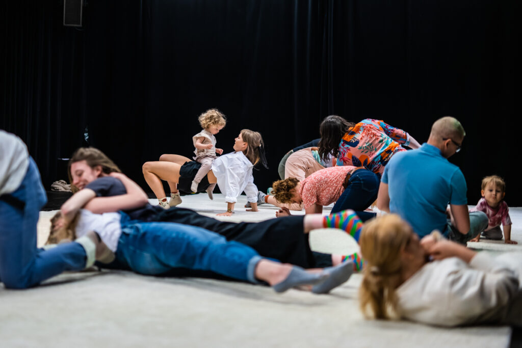 Na scenie grupa dzieci i dorosłych. Na przedzie kilkoro osób lezy. W tle po prawej kilkoro siedzi lub klęczy. Po lewej kobieta robi mostek, na jej brzuchu siedzi dziecko.