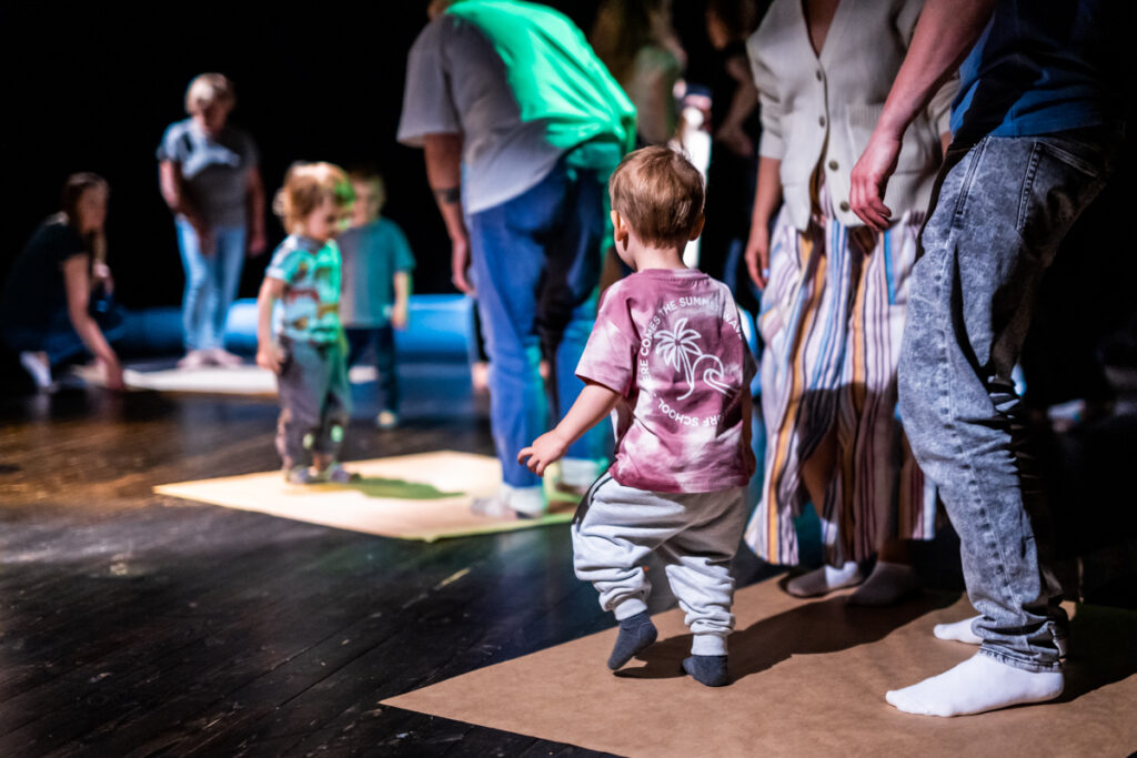 Na scenie rozłożone są kwadratowe maty z papieru. Na których stoi grupa dzieci i dorosłych.