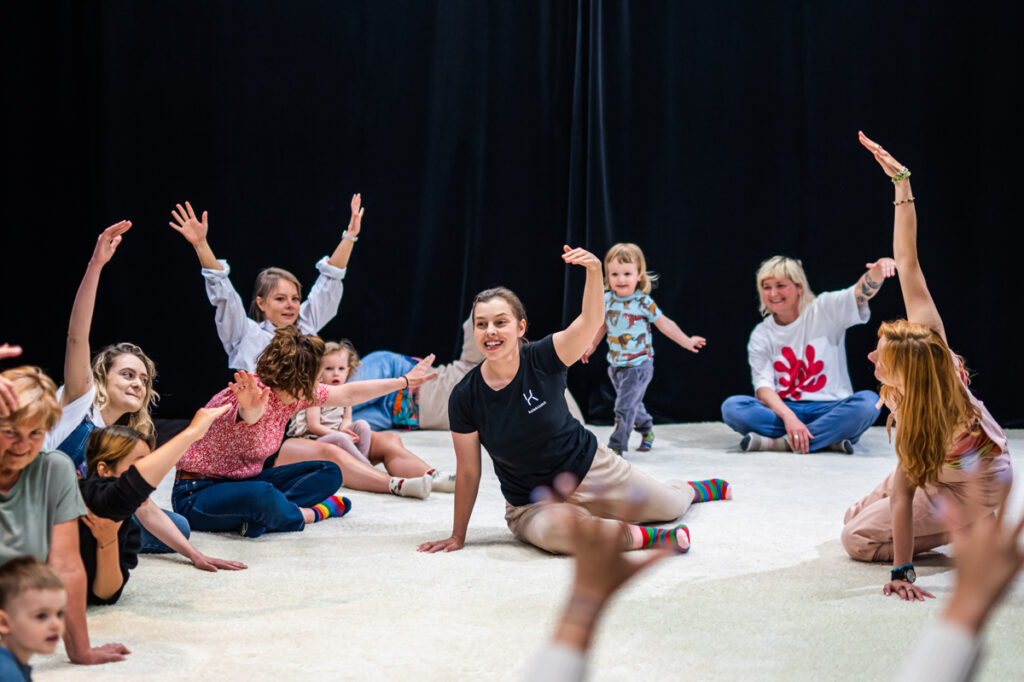 Na scenie grupa dzieci i dorosłych siedzi na podłodze. Unoszą do góry ręce.