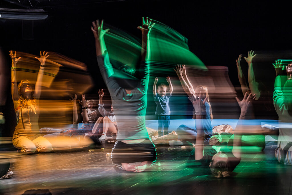 Na scenie grupa dzieci i dorosłych. W różnych pozycjach: część leży, część siedzi, inni stoją. Wszyscy mają wysoko uniesione ręce.