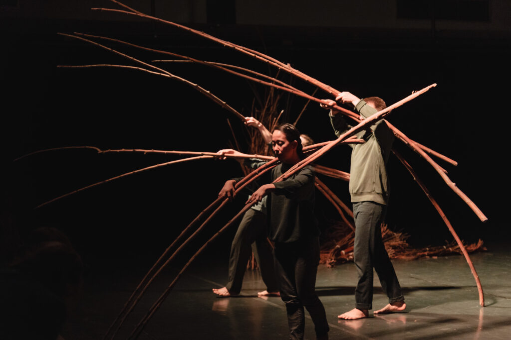 Troje aktorów na scenie. Każdy trzyma na ramionach długie wygięte gałęzie.