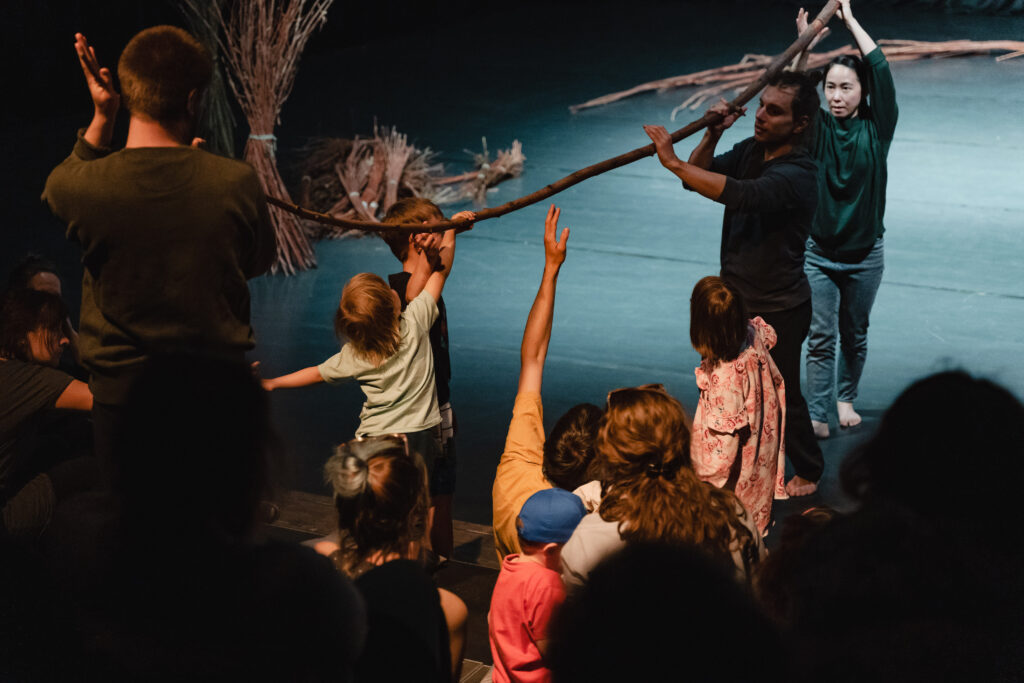 Na scenie. Aktorzy trzymają długą gałąź. Jeden z aktorów stoi pośród widowni. Dzieci z widowni sięgają rękoma do gałęzi.