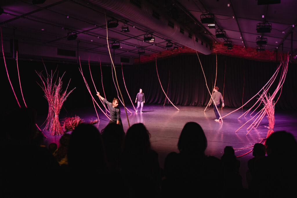 Na scenie trójka aktorów chodzi do około. Na scenie rozstawione są długie gałęzie oparte o podłogę.