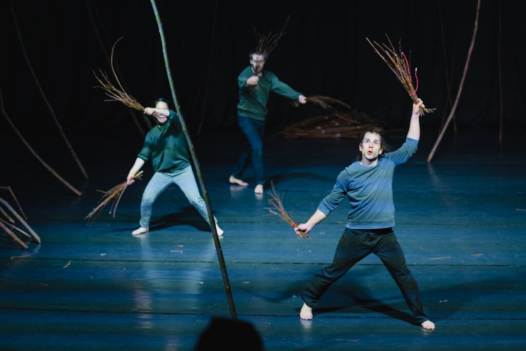 Trójka aktorów na scenie. Mają w rękach pęki gałązek i nimi machają.