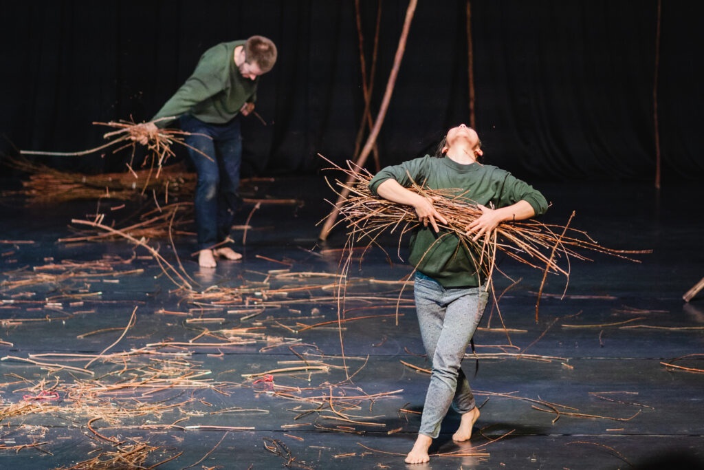 Na scenie dwójka aktorów. Aktorka z przodu trzyma w rękach pęk patyków. Ma mocno odgięta do tyłu głowę. W tle aktor rozrzuca patyki po podłodze.