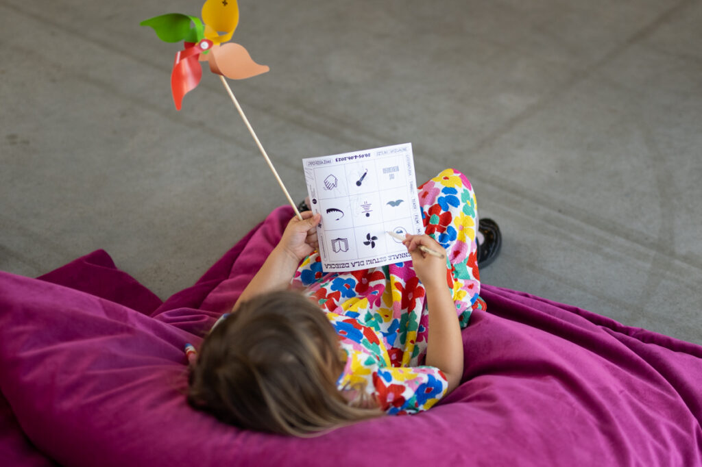 Dziecko leży na wielkiej poduszce. Ma w rękach kartę do gr w bingo i kolorowy wiatrak.