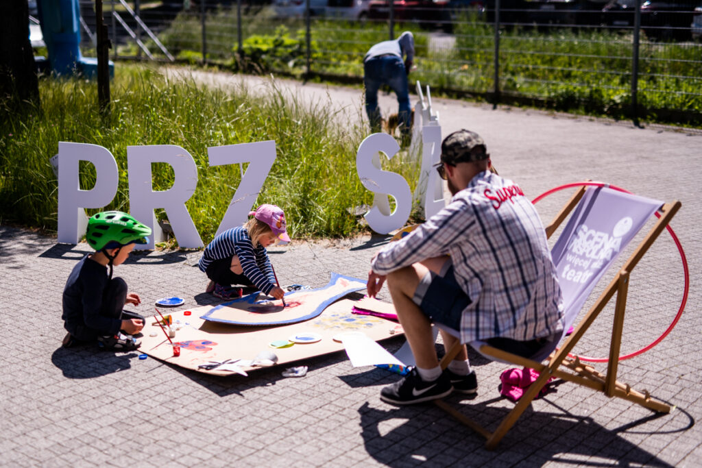 Po prawej mężczyzna siedzi na leżaku. Przed nich dwójka dzieci maluje na wielkim, rozłożonym na ziemi kartonie.